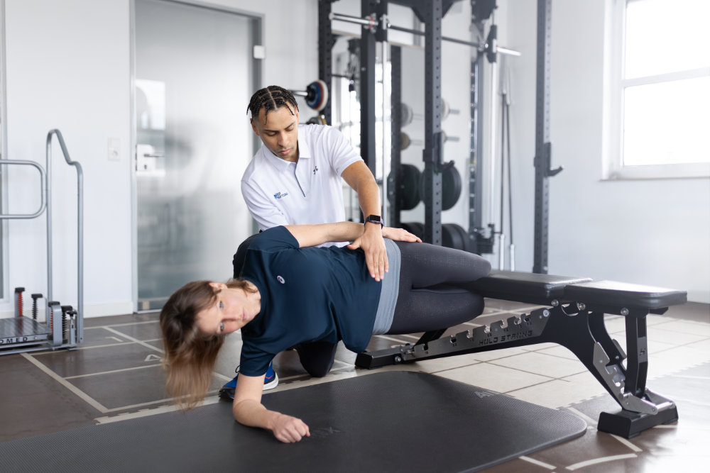 Ein Trainer unterstützt eine Frau bei einer Übung zur Kräftigung und Stabilisierung der Hüfte, während sie in Seitlage auf einer Trainingsbank liegt.